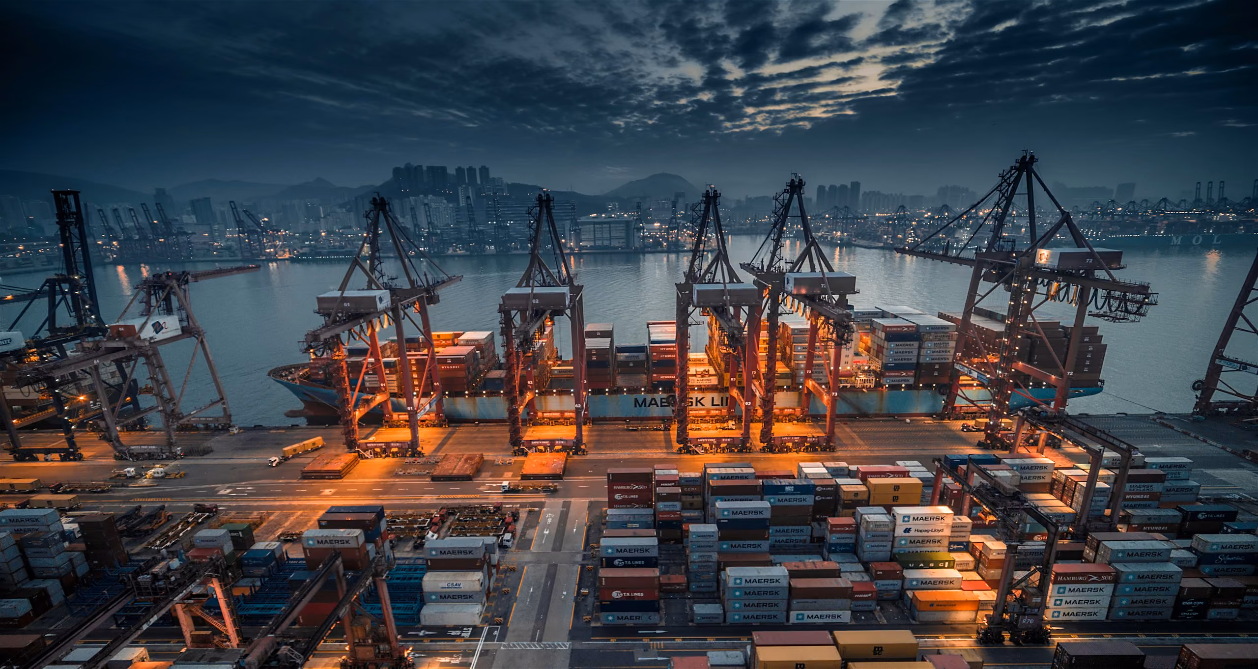 A container ship being loaded by gantry cranes at a busy port at dusk
