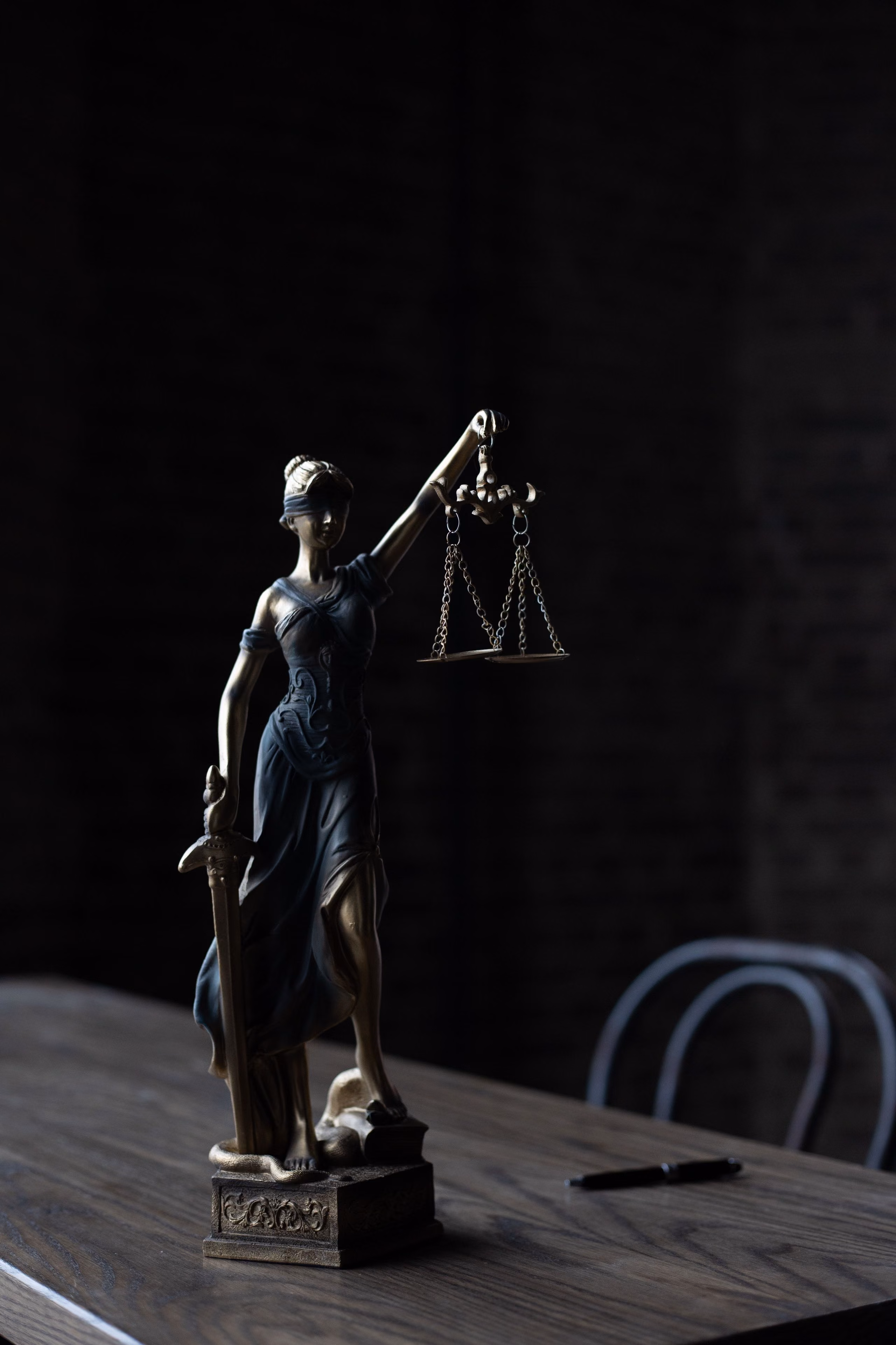 A bronze figure of Justice holding a balance, standing on a wooden table against a dark wall