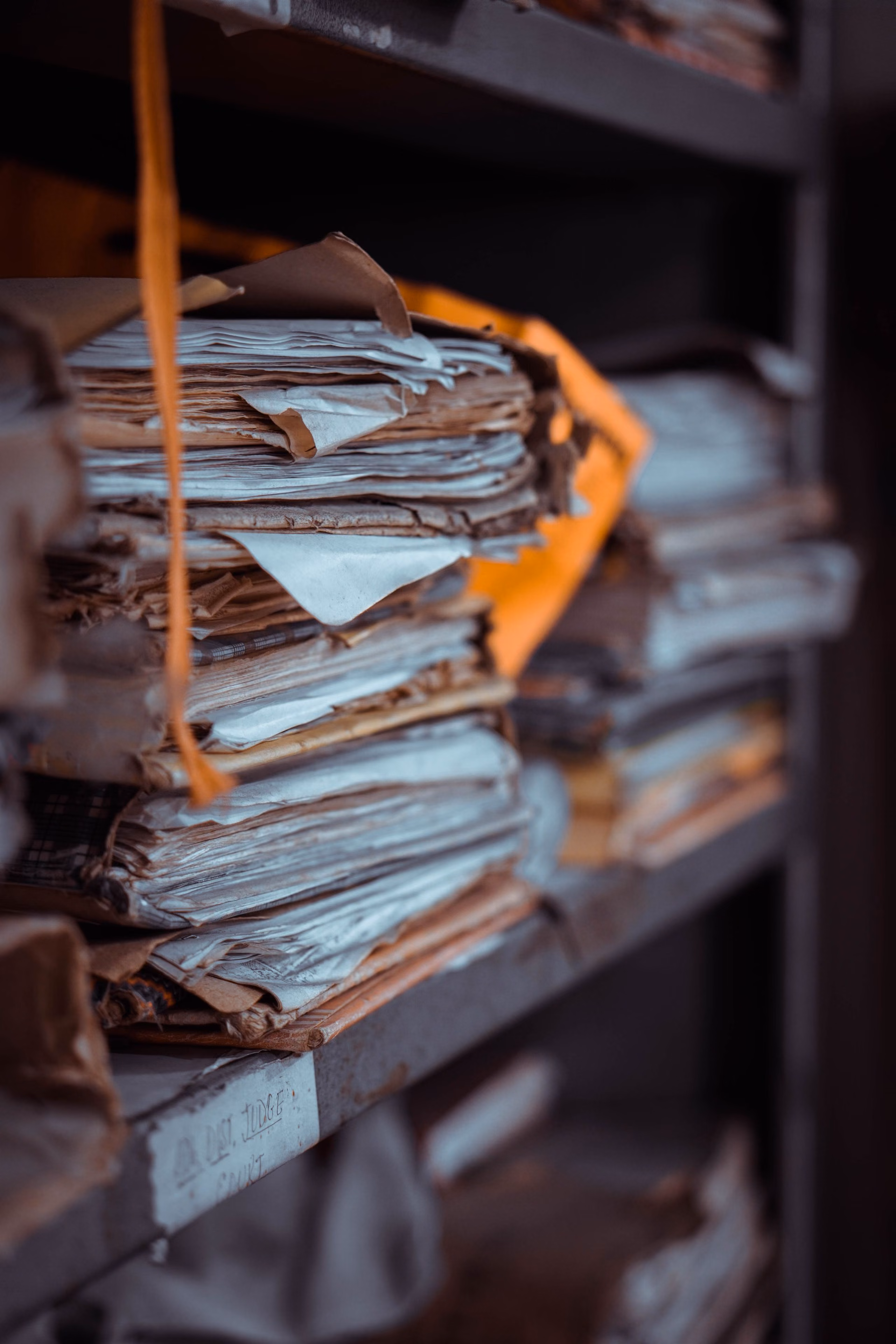 Worn paper files stacked on the metal shelves of a record room, their edges frayed with age